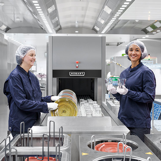 Two employees in an industrial kitchen operating a HOBART dishwashing line loaded with dishes and glasses.