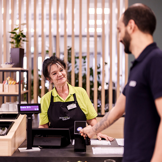 Payment process in a modern company cafeteria: A customer makes a contactless card payment at an electronic POS terminal. The amount is shown on a digital display. Wooden slats and plants in the background create a welcoming atmosphere.