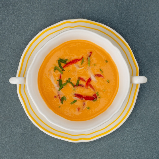 White soup cup with yellow rim on a plate, filled with creamy orange soup. The cup has two handles and is placed on a gray table.