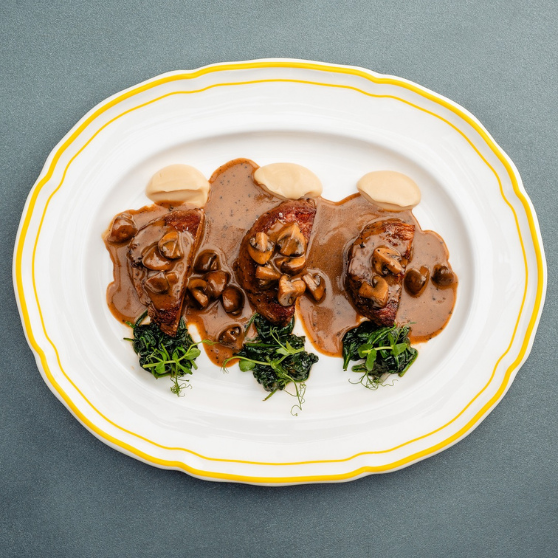 White plate with yellow rim featuring three pieces of meat with mushroom cream sauce, garnished with mushrooms, light puree and green herbs, elegantly plated on a gray table.
