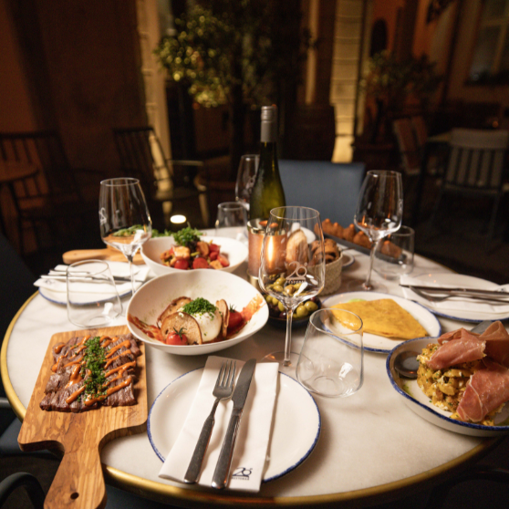 Beautifully set outdoor restaurant table with several dishes, appetizers, bread, a bottle of wine and multiple glasses arranged for a relaxed dining experience.