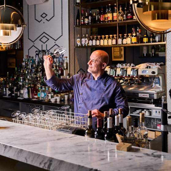 Person behind a bar examining a clean glass, with a glass rack on the counter, bottles arranged in the foreground and a fully stocked back bar with a professional coffee machine in the background.