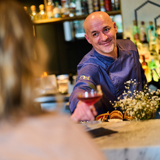 Bartender serving a drink across the bar counter to a guest, with bottles and bar shelves visible in the background.