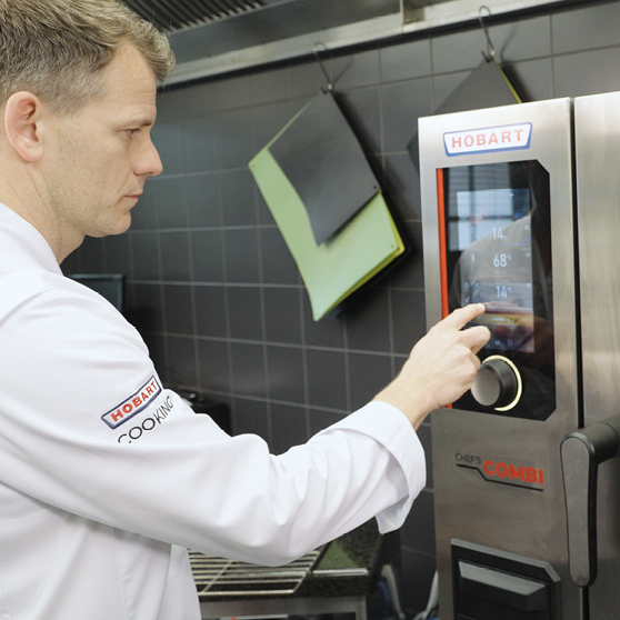 Chef operating the touchscreen control panel of a HOBART CHEF’S COMBI in a professional kitchen, adjusting settings for precise and efficient cooking workflows.