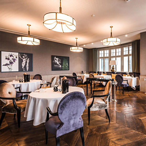 Elegant fine‑dining restaurant interior featuring round tables with white linens, upholstered chairs, artistic wall decor, and warm lighting creating an exclusive atmosphere.