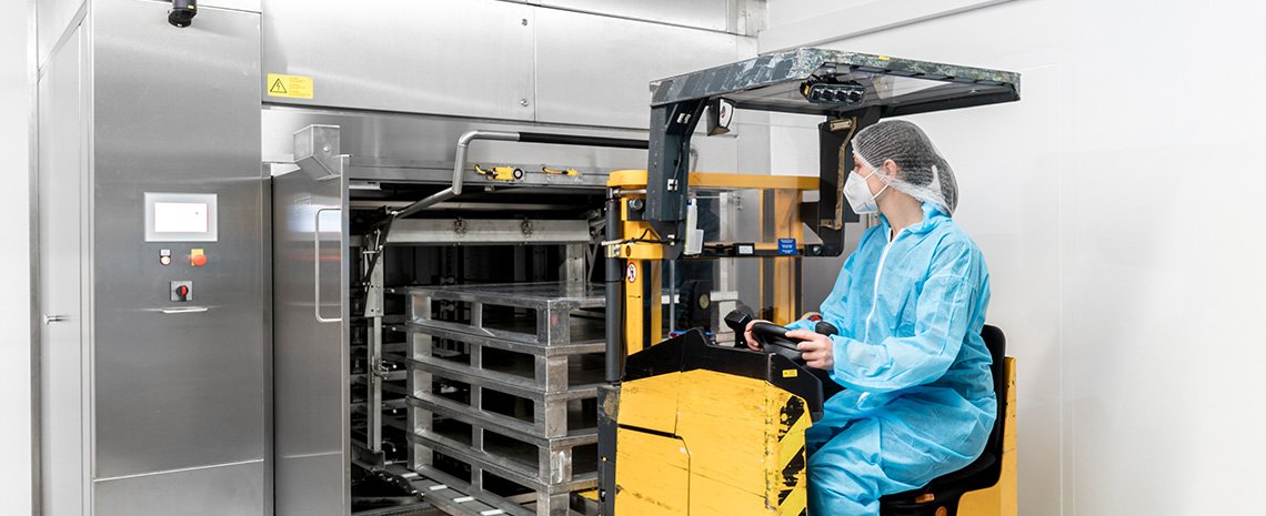A person in protective clothing operating a yellow lift vehicle in front of a stainless‑steel HOBART pallet cleaning system with stacked pallets positioned inside the machine.