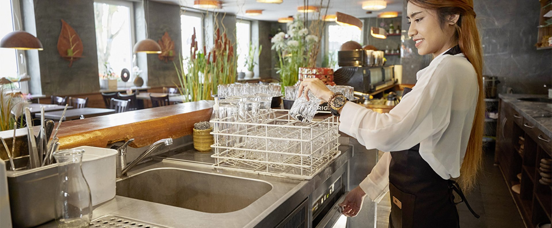 A person is working behind the counter of a modern restaurant, placing clean glasses into a rack above a HOBART glasswasher to ensure an efficient and hygienic workflow in the beverage service area.