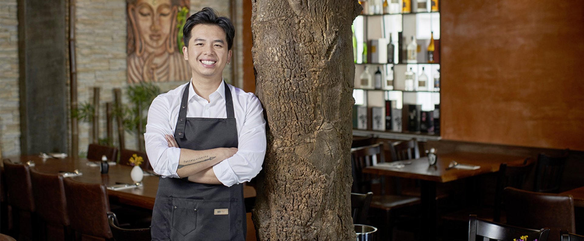 A person wearing a service apron is standing inside a stylish restaurant interior, positioned near a decorative tree element, representing the atmosphere and concept of the dining establishment.