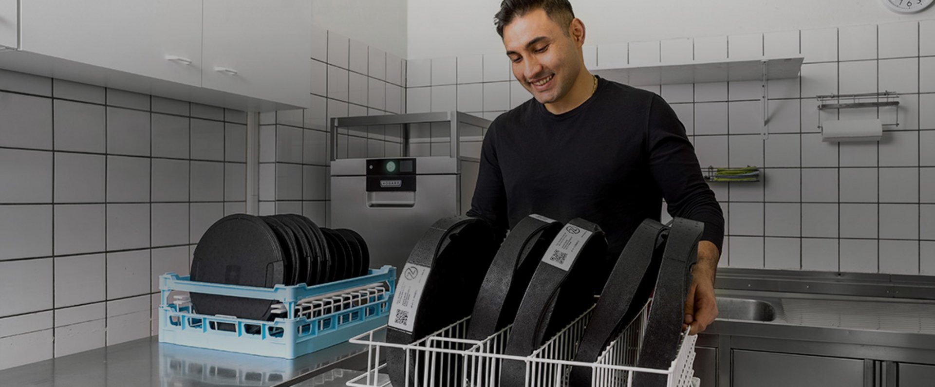 A person loads reusable pizza boxes from rezzeat into a dish rack inside a tiled commercial kitchen, preparing them for hygienic cleaning in a professional dishwashing process supported by HOBART technology.