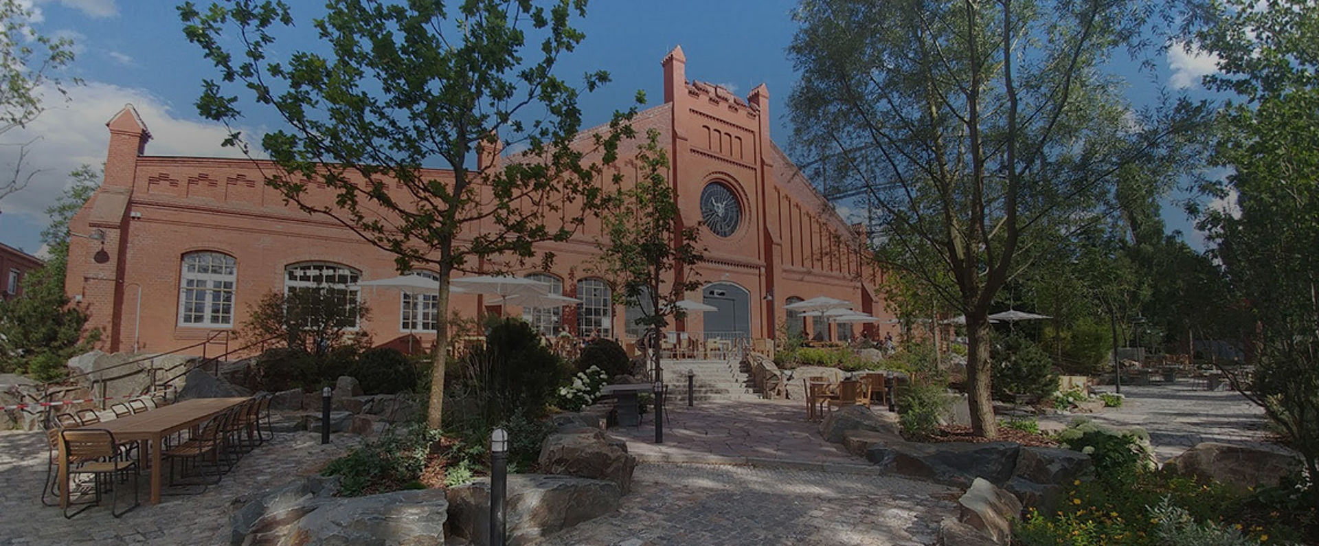Exterior view of the Stone Brewing Berlin location showing the historic red‑brick building, spacious outdoor seating areas, trees, and landscaped pathways that create a welcoming atmosphere for guests.