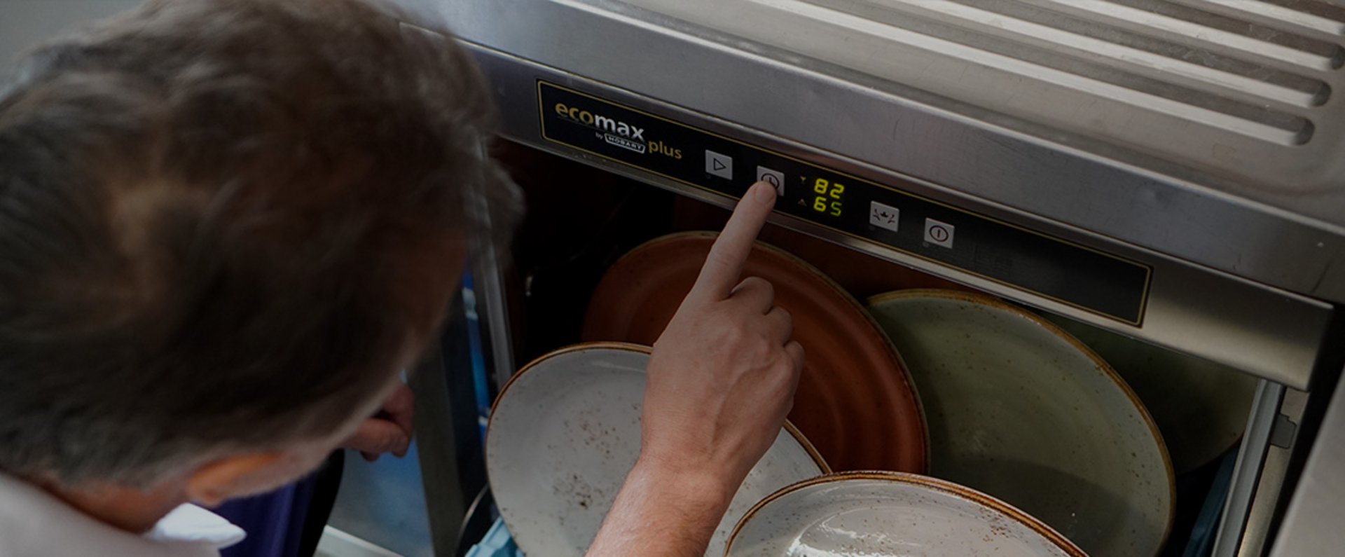 In a professional kitchen, the cook operates an ecomax plus undercounter dishwasher by pressing a button on the control panel above a loaded rack filled with ceramic plates, illustrating an efficient workflow during daily dishwashing operations.
