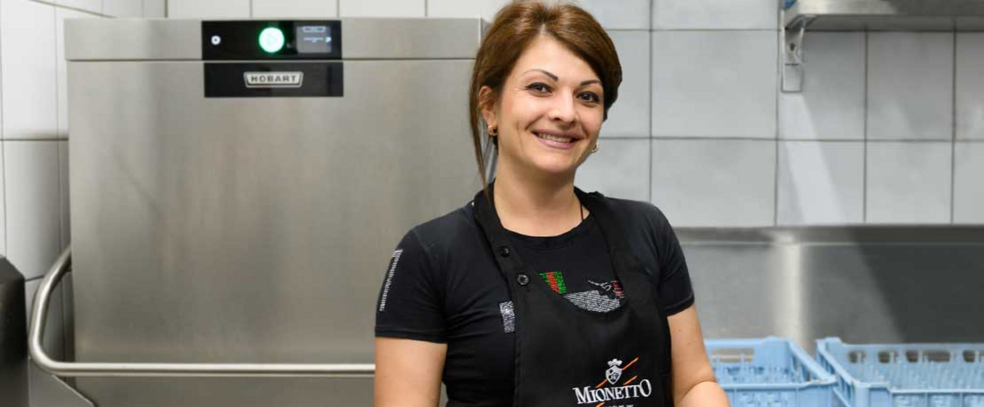 Staff member standing in front of a HOBART commercial dishwasher inside a tiled kitchen, with dish racks and stainless steel workspace arranged around the machine.