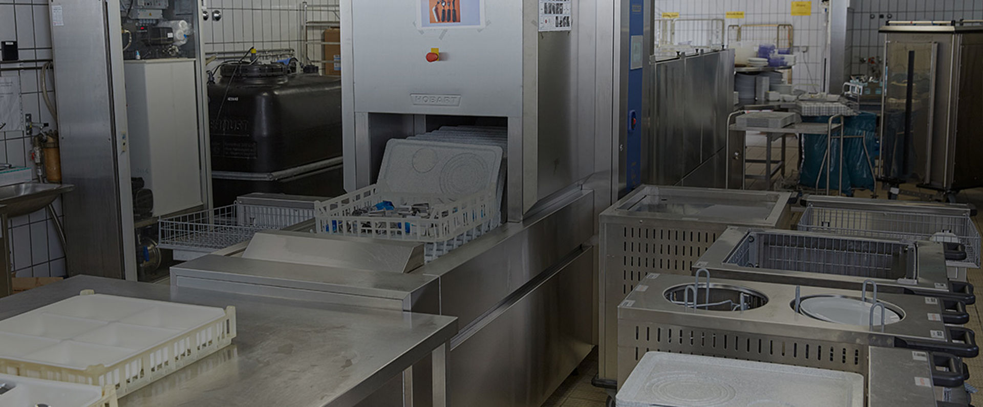 Professional dishwashing area featuring a HOBART conveyor dishwasher in continuous operation, surrounded by preparation stations and equipment designed for efficient cleaning of large quantities of trays and tableware in a hospital environment.
