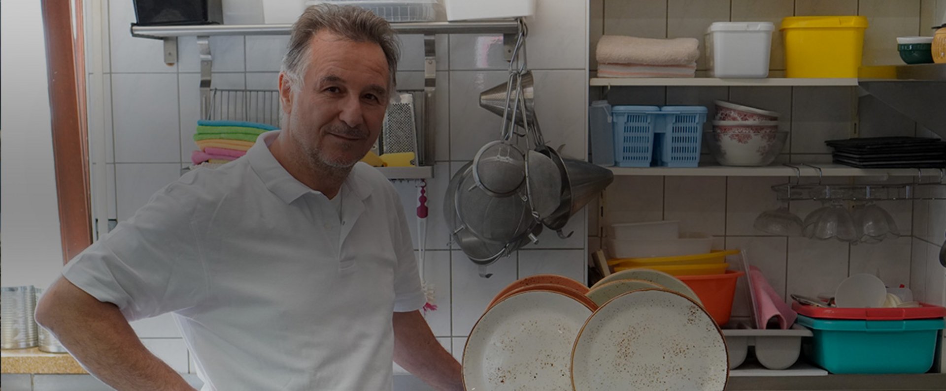 In the open kitchen of the Zafferano restaurant, the cook stands beside stacked ceramic plates in front of a stainless‑steel work area, surrounded by neatly arranged kitchen tools, strainers, containers, and shelving, giving an authentic impression of the restaurant’s daily workflow.