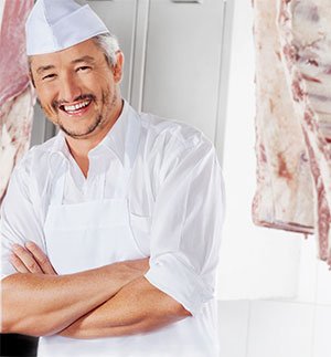 Person wearing white kitchen clothing and a white cap standing in a room with hanging meat in the background.