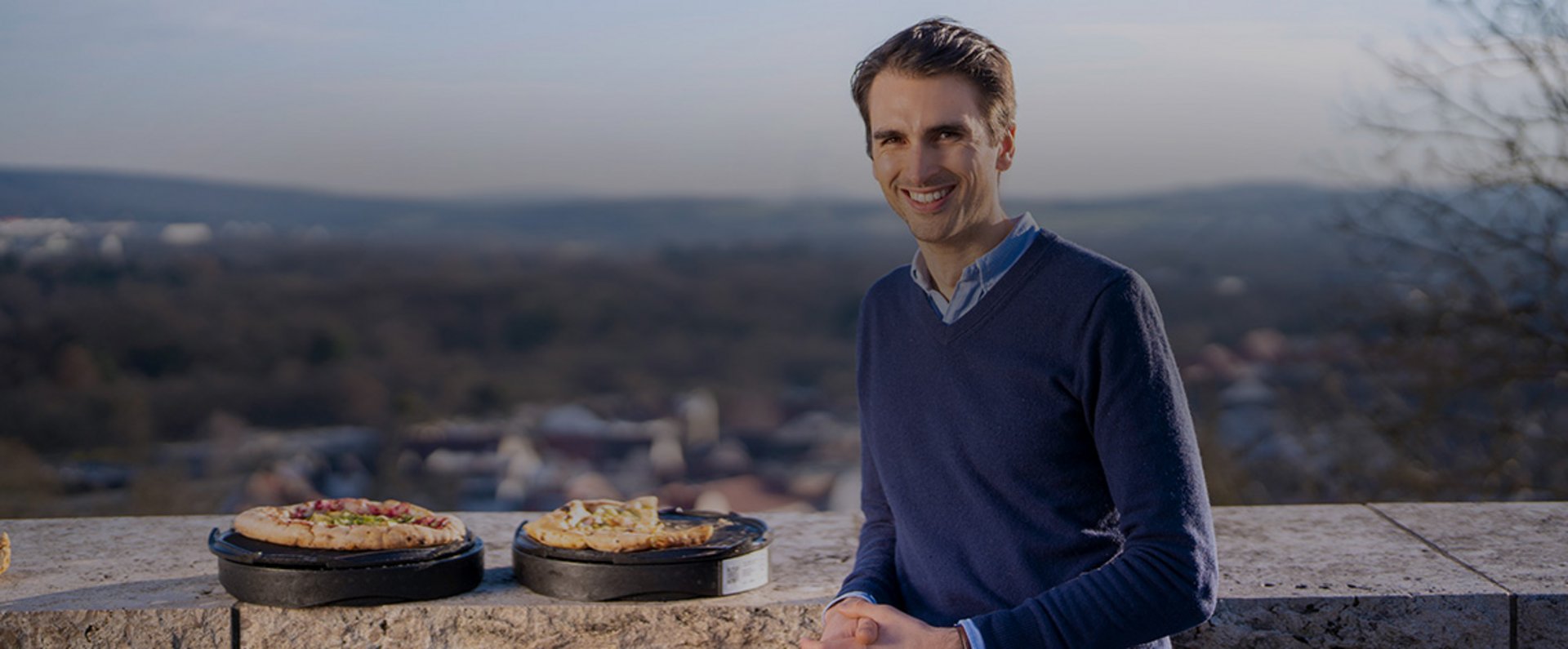 A person sits outdoors with reusable rezzeat pizza boxes placed on a stone railing, each holding a freshly prepared pizza, with a scenic view of the town and landscape in the background.