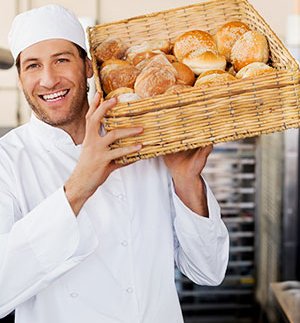 Person in a white kitchen outfit holding a large wicker basket filled with bread rolls in a commercial kitchen setting.