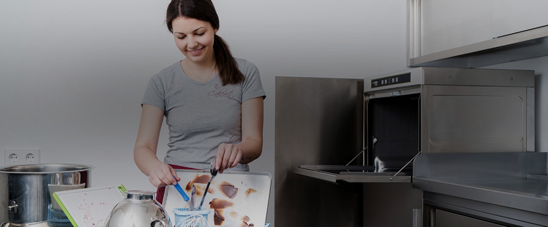 A person prepares ingredients on a stainless‑steel work surface in a production kitchen equipped with an open ecomax by HOBART undercounter dishwasher and various baking tools and mixing bowls.