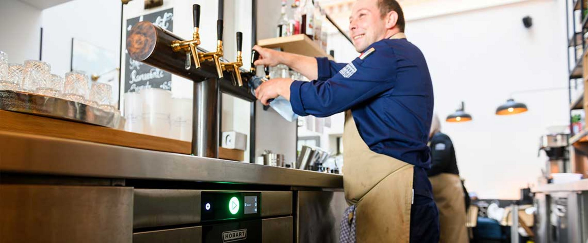 Staff member using a beverage tap station above a HOBART undercounter dishwasher in a bar area equipped with glasses, bottles, and stainless steel work surfaces.