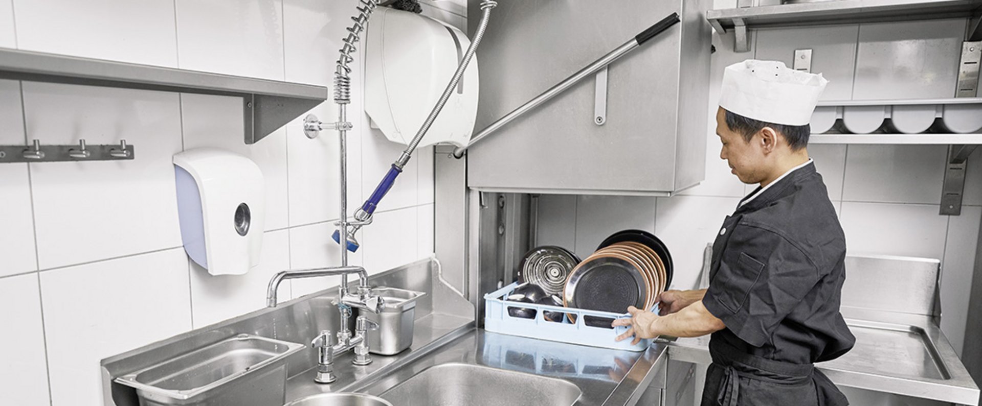 A person is working in a professional kitchen, loading a rack filled with pots and pans into a commercial dishwasher to support an efficient and hygienic cleaning process in the back-of-house area.
