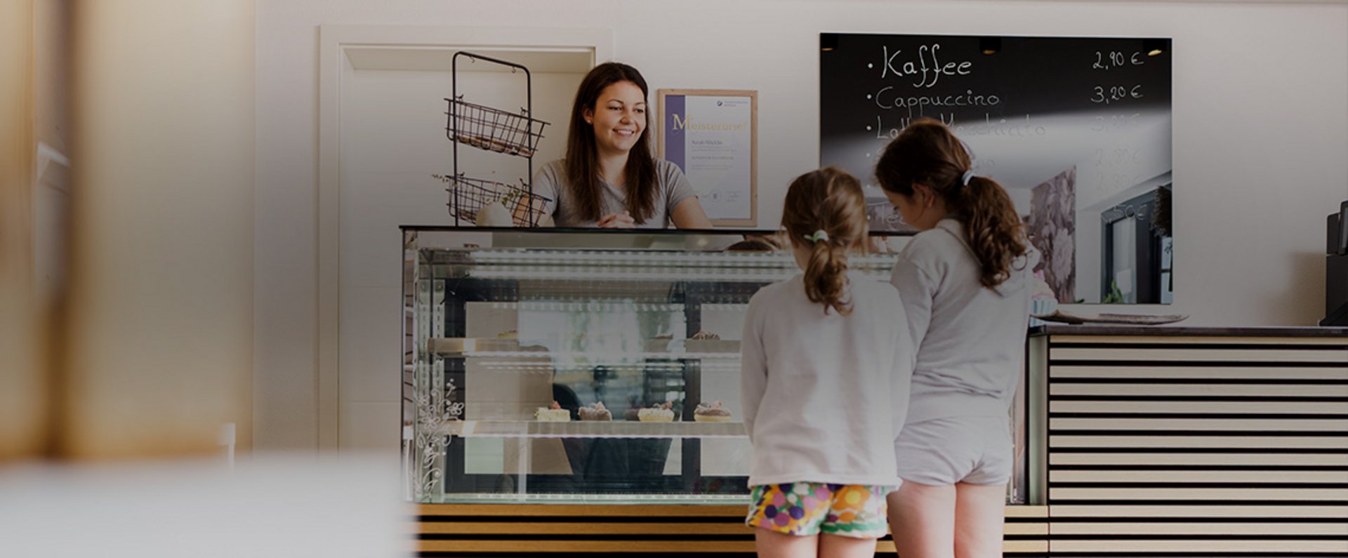A person stands behind a pastry counter in a bright shop interior while two children look at the desserts displayed in the glass case, with various pastries and cake items arranged for sale.