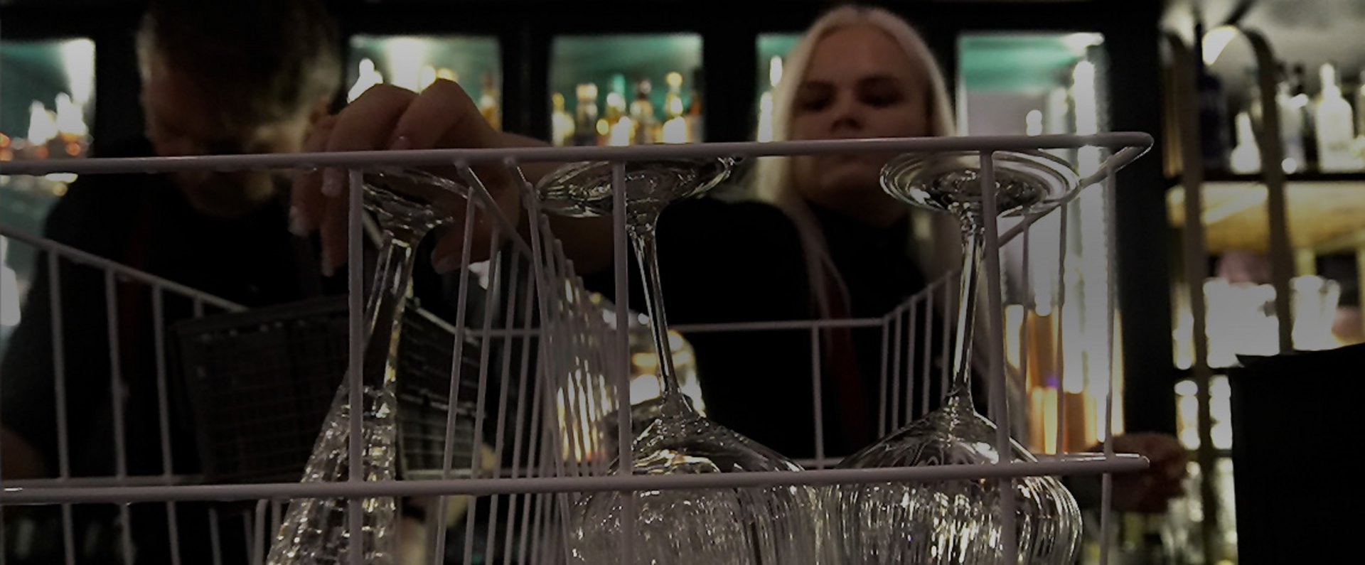 Freshly washed glassware in a bar rack positioned on the counter, with staff working behind the rack in a dimly lit bar environment equipped for efficient glass handling.