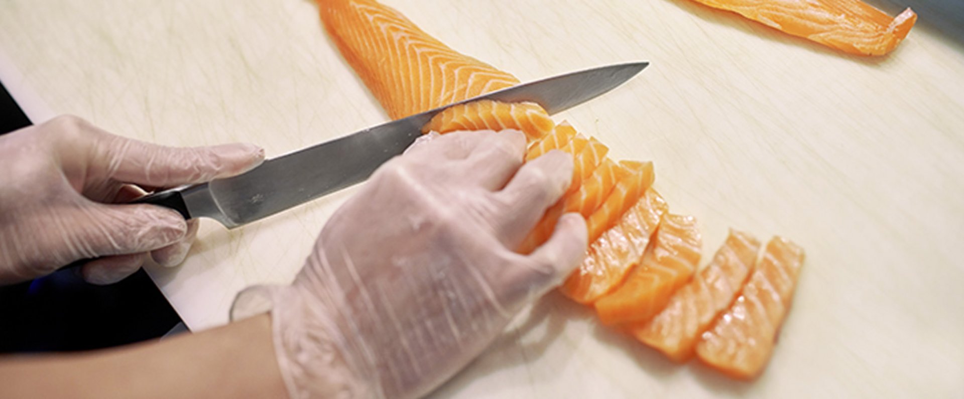 Hands wearing protective gloves are slicing fresh salmon on a cutting board as part of professional food preparation in a commercial kitchen.