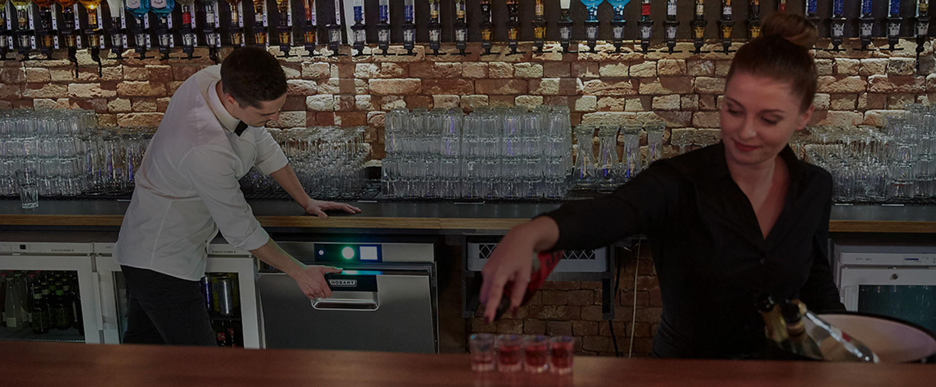 In a lively nightlife bar, one staff member serves shots at the counter while another operates a HOBART glasswasher beneath a long row of polished glasses and illuminated bottle racks set against a brick wall, illustrating an efficient and hygiene‑focused workflow during busy bar service.
