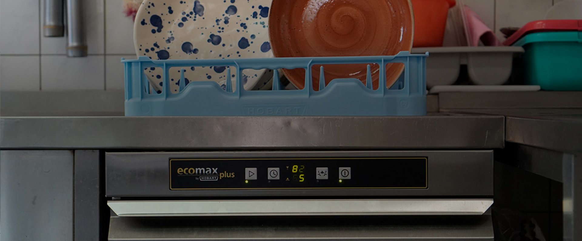 A HOBART ecomax plus undercounter dishwasher is shown in a professional kitchen environment with a dish rack placed on top, holding patterned and colored plates, highlighting the compact and efficient setup for everyday dishwashing operations.