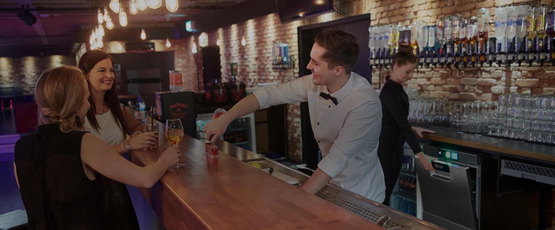 In a stylish nightlife bar, a bartender serves drinks to guests at the counter while another staff member works near a HOBART glasswasher, with illuminated bottle racks and neatly arranged glassware along the brick wall, illustrating a fast and hygiene‑focused workflow during busy club operations.