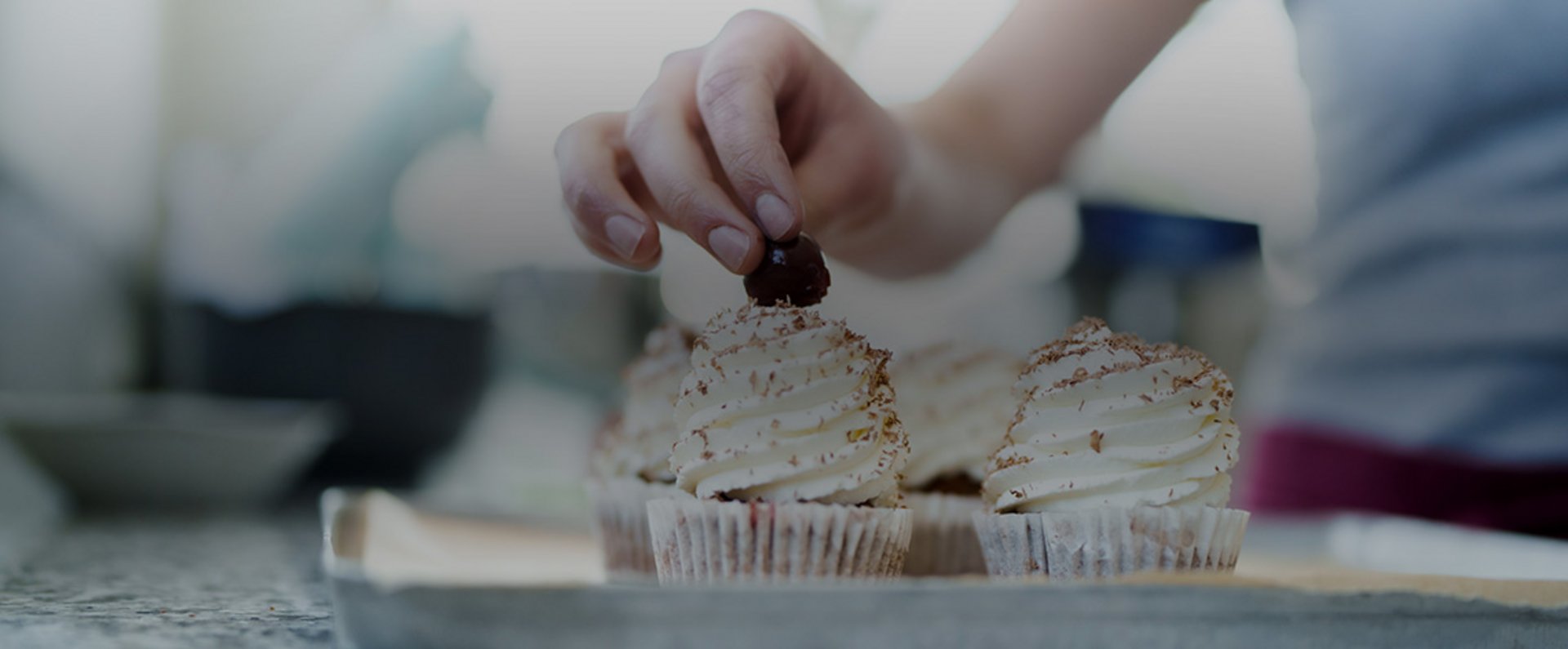A hand places a decorative element onto freshly piped cupcakes arranged on a baking tray, highlighting the detailed finishing work in the pastry preparation process.