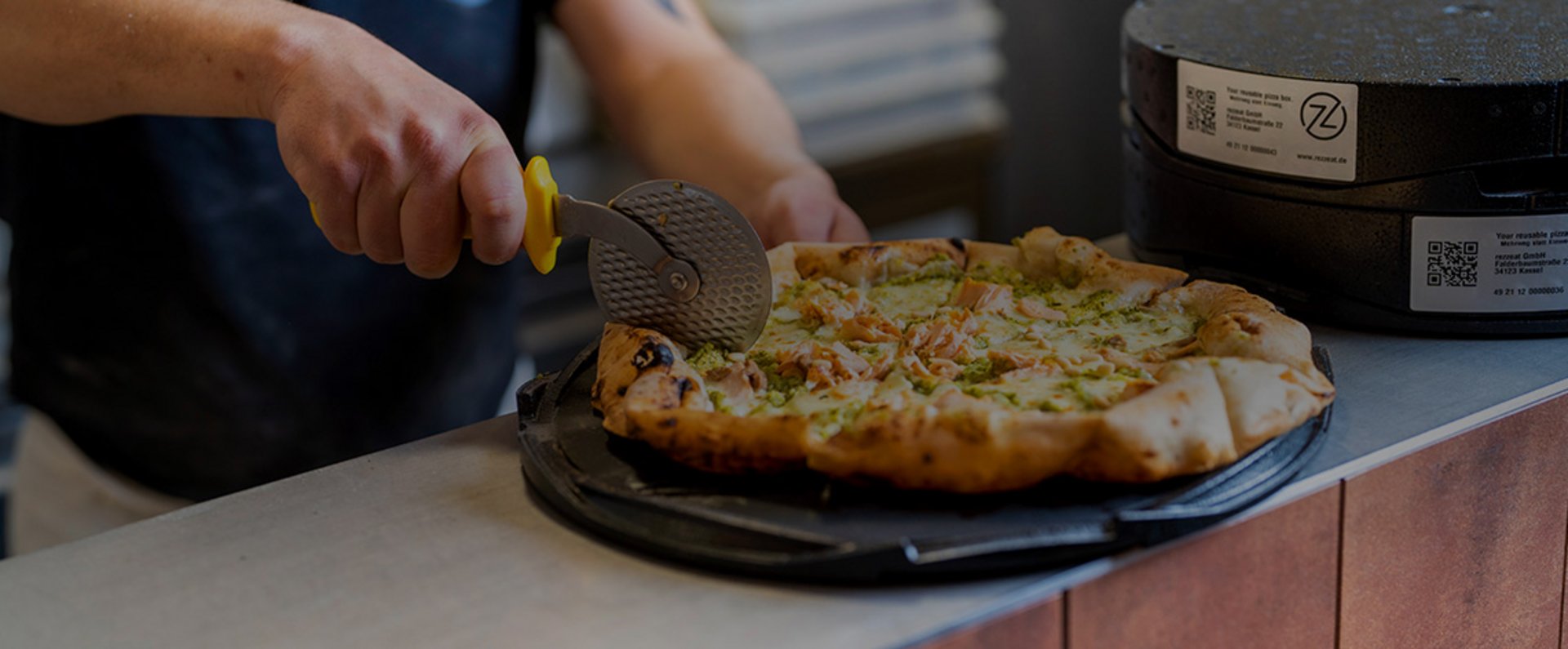 A person uses a pizza cutter to slice a freshly baked pizza placed on the reusable rezzeat pizza box base, showing the preparation process before sustainable takeaway transport.