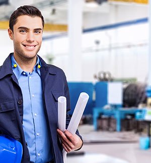 Person wearing a dark blue work jacket holding rolled‑up plans in an industrial workshop with machinery in the background.