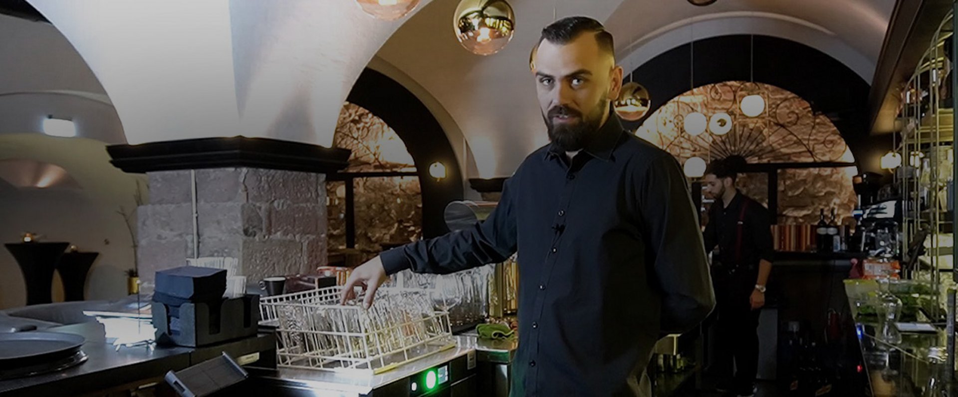Staff member standing in a vaulted bar interior next to an open HOBART undercounter dishwasher filled with glass racks, demonstrating efficient glasswashing operations.
