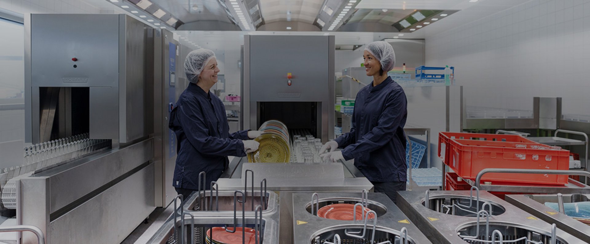 Two staff members working at a HOBART conveyor dishwashing system, loading racks of plates into the organized high‑efficiency cleaning workflow in the MTU Aero Engines commercial kitchen.