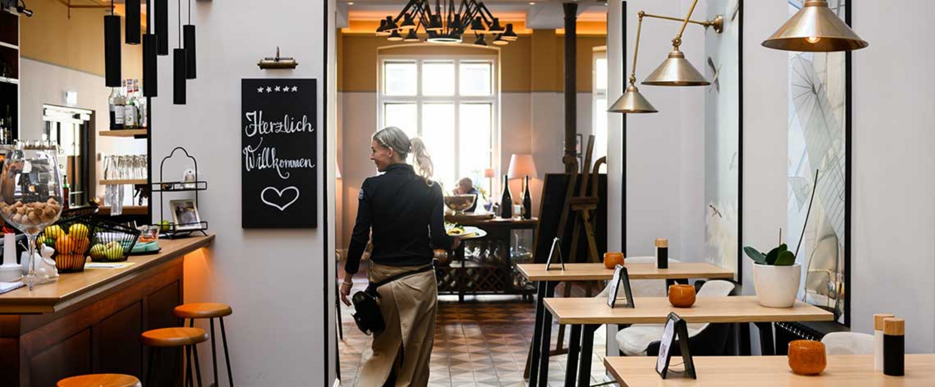 Interior view of the restaurant at Anklamer Hof showing wooden tables, modern lighting, a bar counter, and a staff member walking through the dining area.