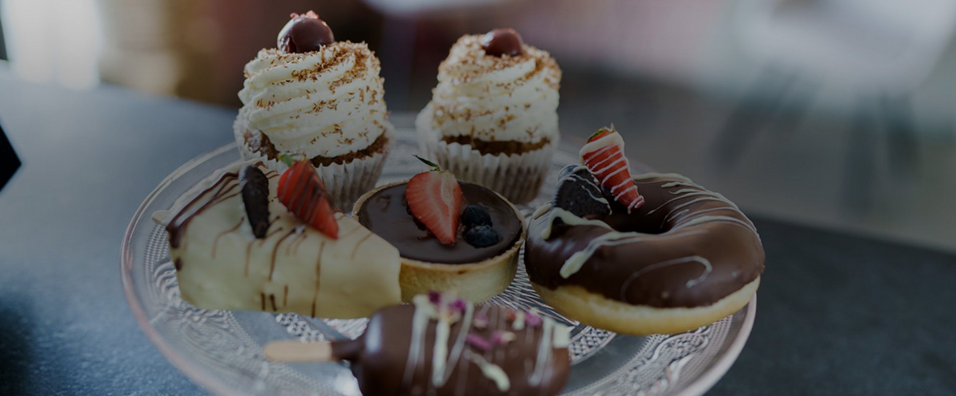 Close‑up view of assorted pastries including cupcakes, donuts, and chocolate‑coated treats arranged on a glass plate, showcasing decorated sweets from Cakeart by Sarah.