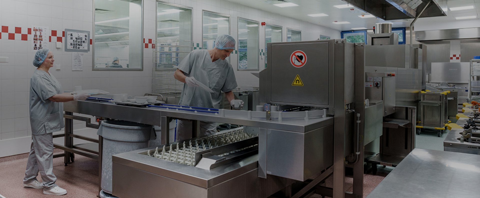 Two individuals work along the loading section of the HOBART autoLINE dishwashing system in the Garmisch‑Partenkirchen hospital kitchen, placing trays and dishware onto the conveyor setup in a brightly lit hygienic workspace.