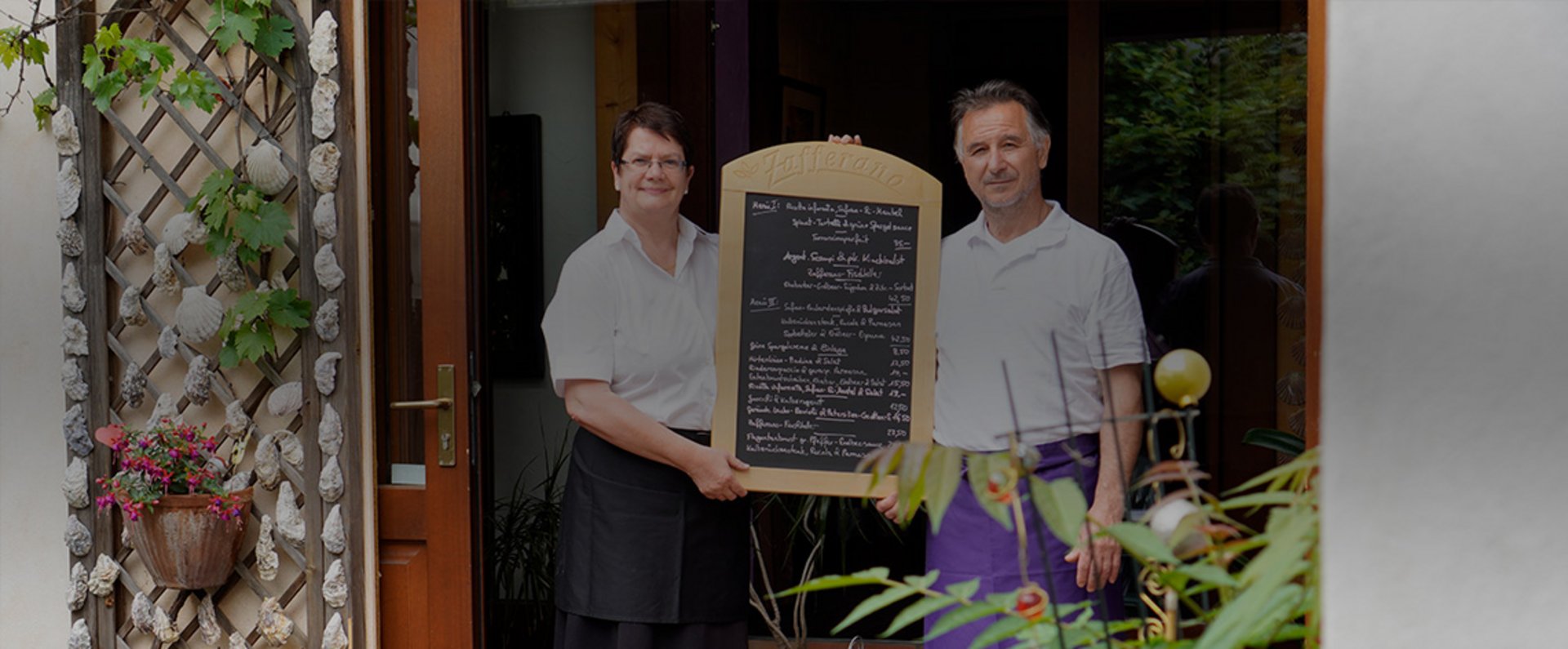 At the entrance of a cozy, Italian‑inspired restaurant, two staff members hold a large handwritten menu board featuring the day’s dishes, highlighting the welcoming atmosphere and the personal, creative approach to fresh cuisine.