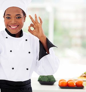 Person wearing a white double‑breasted kitchen jacket and chef hat making an OK hand gesture in a bright kitchen setting with food items in the background.