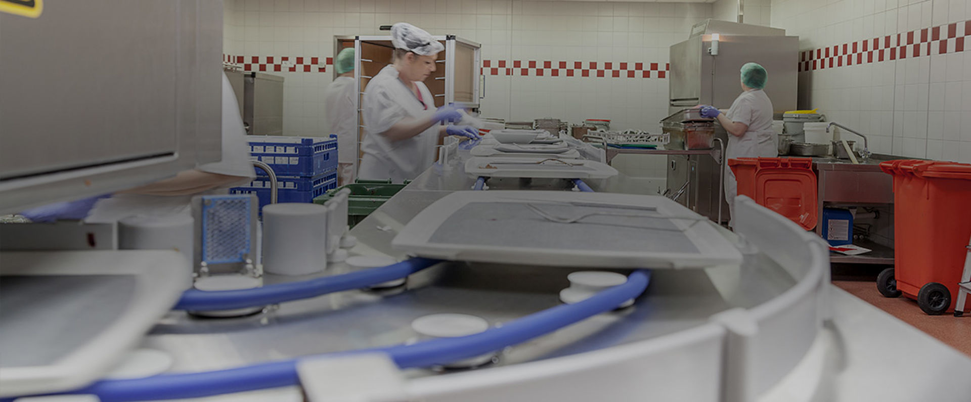 Conveyor section of the HOBART AutoLINE system in the SHG Clinic Völklingen, showing trays moving along the automated track while staff members work at various stations in the dishwashing area.